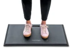 En person iført lyserøde sneakers står på en sort anti-træthedsmåtte med en udskæring i håndtaget. Mousetrapper Active Ståmåtte, mærket nær den nederste kant, komplementerer deres sorte bukser og giver stil og komfort. - Ergoshop - Ergonomiske kontormøbler til erhverv
